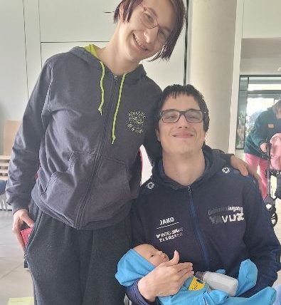Mann hat ein Simulationsbaby in der Hand. Er sitzt in einem Rollstuhl. Seine Partnerin steht nehmen ih. Ihre linke Hand liegt auf seiner linken Schulter. Beide lächeln zufrieden.